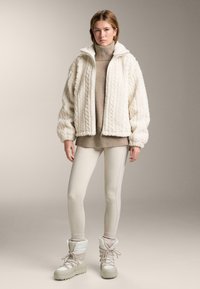 Mjuk vit zip-up jacka med hög krage, kombinerad med en beige polotröja, krämfärgade leggings och vita snörstövlar.