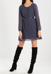 Žena v modré šaty ke kolenům s průsvitnými dlouhými rukávy, černým páskem a černými sukovými kozačkami po kolena.