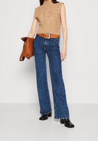 Nainen, jolla on päällä vaaleanbeige ilmanhihaa oleva neulepaita, ruskea vyö, siniset leveälahkeiset farkut ja mustat saappaat, pitää isoa ruskeaa nahkakäsilaukkua sivussa.