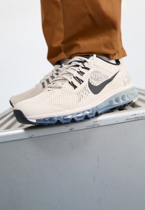 Baskets Nike Air blanches avec un swoosh noir et des semelles à coussin d’air transparentes, portées avec un pantalon marron sur une surface métallique.