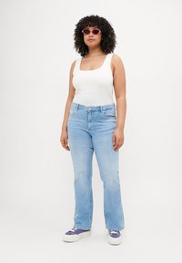 White tank top, light blue flared jeans, and purple sneakers. Round sunglasses and curly hair. Simple design with a casual fit.