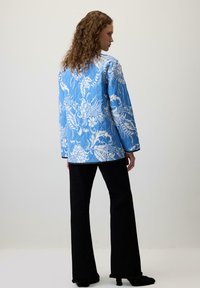 Pamučena plava jakna s bijelim cvjetnim uzorcima, dugim rukavima i crnim rubovima, u kombinaciji s širokim crnim hlačama. Glatka tekstura, opušteni kroj.