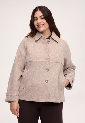 Woman with long dark hair smiles wearing a beige buttoned jacket with subtle swirl patterns and dark pants against a plain background.