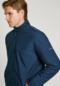 Marinblå jacka gjord av ett slätt, lätt tyg med hög krage, framdragkedja och diskret logotypdetalj på ärmen.