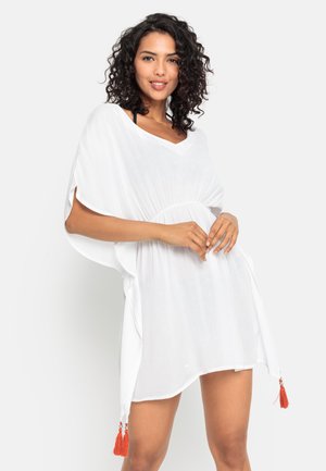 Uśmiechnięta kobieta z kręconymi włosami ma na sobie luźną białą sukienkę z pomarańczowymi frędzlami, pozując na jednolitym jasnym tle.