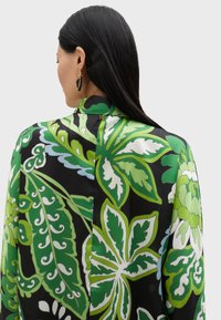 Svart blus med stora gröna blommönster och vita accenter, med hög krage och guld detaljer på baksidan.