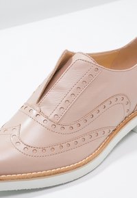 Chaussure en cuir rose clair avec des détails perforés et des accents ondulés. Elle présente un design à enfiler et une semelle blanche contrastante.