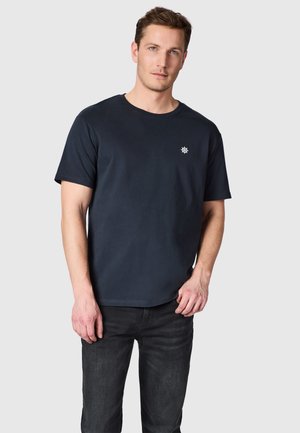 Man die een zwart T-shirt met korte mouwen draagt met een klein wit stuurwiel-logo op de borst en donkere spijkerbroek, staand tegen een lichtgrijze achtergrond.