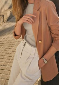 Jasnobrązowa marynarka na szarym topie, w połączeniu z kremowymi spodniami z wysokim stanem. Marynarka ma złoty guzik i dopasowane rękawy, na nadgarstku widoczny zegarek.