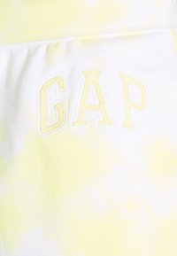 Tissu blanc avec un motif tie-dye jaune. Présente une broderie jaune en relief du mot "GAP" en police épaisse. Texture douce, design décontracté.