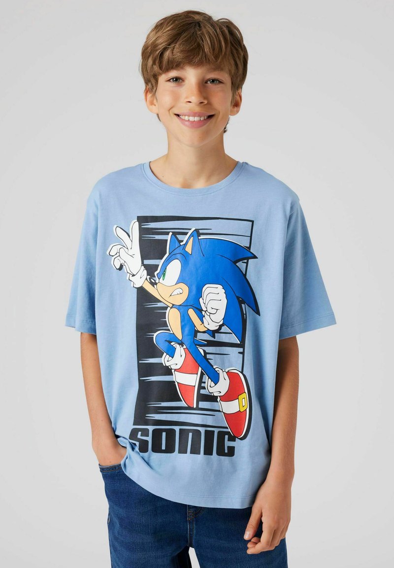 Băiat zâmbitor purtând un tricou albastru deschis cu Sonic the Hedgehog și blugi albaștri, stând pe un fundal gri simplu.