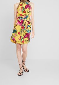 Woman wearing a yellow sleeveless halter dress with pink and green tropical floral print and black lace-up flat sandals.