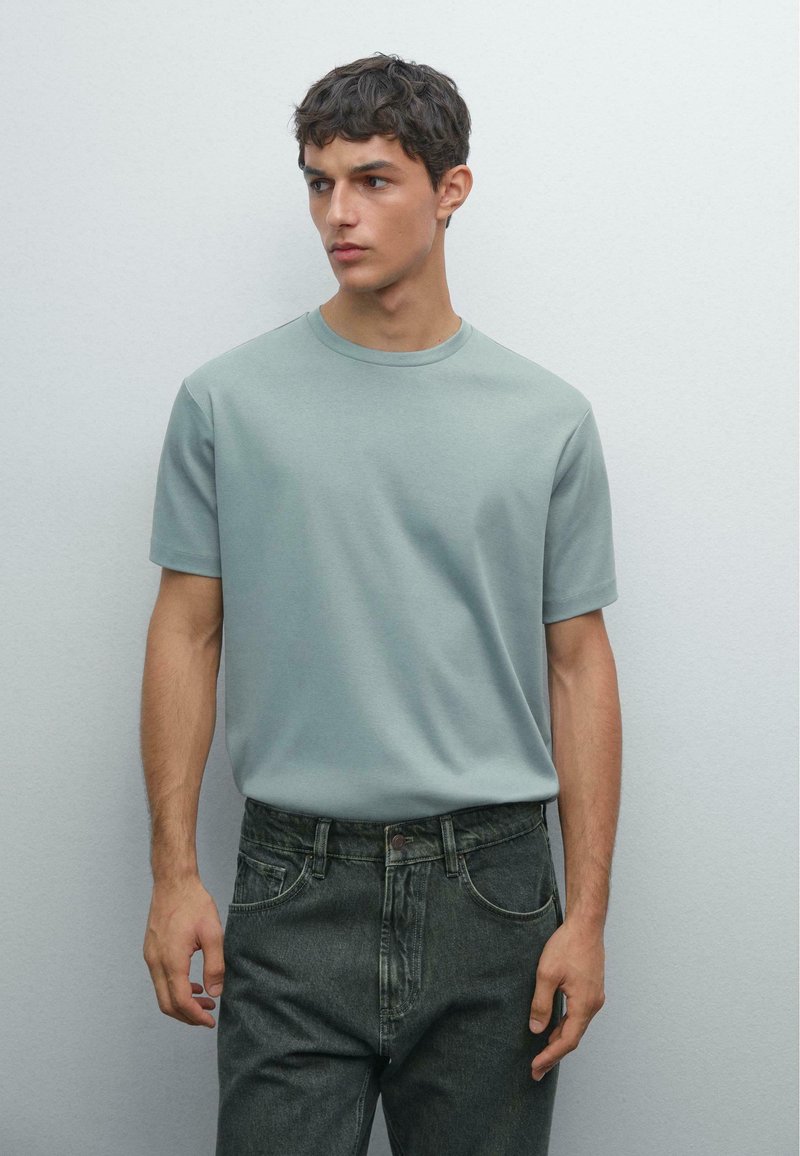 Ung man med kort mörkt hår, iklädd en ljusblå-grön kortärmad t-shirt och mörkgrå jeans, stående mot en enkel ljus vägg.