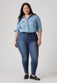 Camisa de denim azul claro com dois bolsos no peito, mangas enroladas; combinada com jeans skinny azul escuro. Mocassins pretos completam o visual.