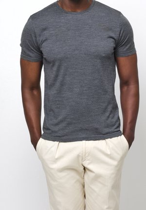 Homme à la peau foncée portant un t-shirt gris chiné ajusté à manches courtes et un pantalon beige clair, les mains dans les poches.
