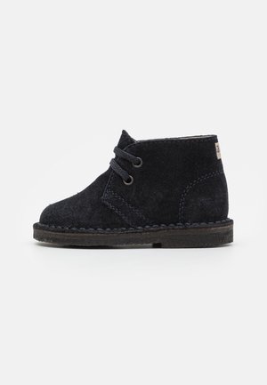 Bottine enfant en daim noir avec deux œillets et lacets noirs, semelle cousue et petite étiquette beige sur le col du talon.