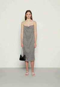 Jovem mulher vestindo um vestido midi sem mangas preto e branco de bolinhas, sandálias brancas, segurando uma bolsa preta com uma alça de corrente prateada.