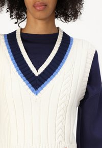 Cream knitted vest with a deep navy V-shaped neckline, featuring blue and navy stripes and cable knit detailing along the sides.