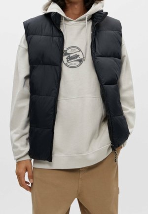 Personne portant un sweat à capuche beige avec un motif rond et un gilet matelassé noir, associé à un pantalon marron clair, debout sur un fond uni.
