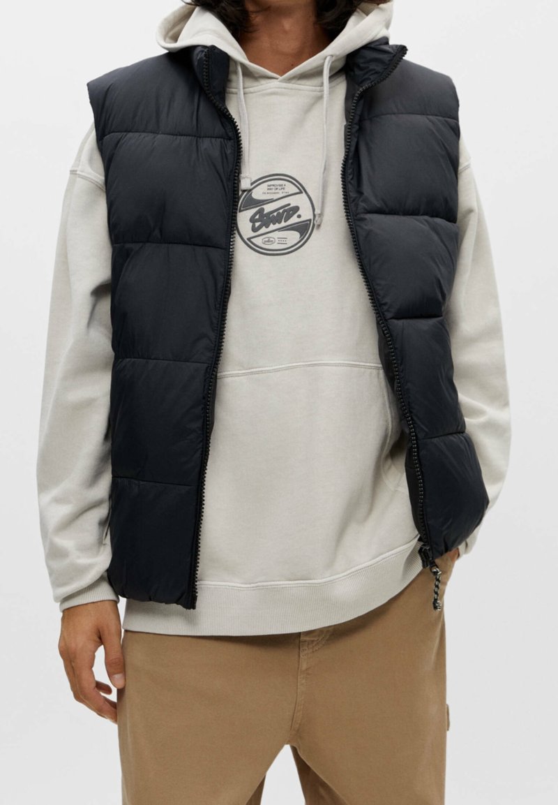 Personne portant un sweat à capuche beige avec un motif rond et un gilet matelassé noir, associé à un pantalon marron clair, debout sur un fond uni.