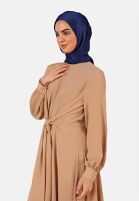 Long beige dress with a front knot feature, long sleeves, and gathered cuffs. Accompanied by a shiny blue hijab. Smooth fabric texture.