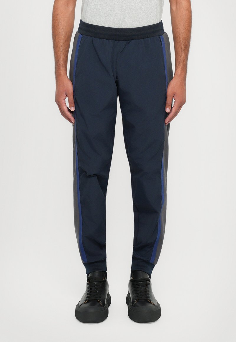 Jūras spēku sporta bikses ir aprīkotas ar elastīgu jostasvietu, sašaurinātu piegriezumu, sānu svītrām tumši zilā un pelēkā krāsā, un gludu, vieglu audumu.