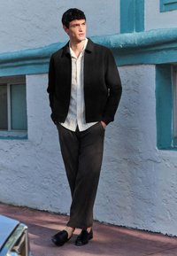 Melna fakturēta jaka, balta krekla apkakle, tumšas vaļīgas bikses un melni ielocīti apavi. Stāv pret gaiši zilu sienu.