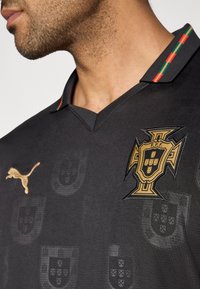 Must must jalgpallimärk, kus on kullaturundusega Portugal vapp ja Puma logo. Krae on punaste, roheliste ja kollaste aktsentidega ning tekstureeritud disainiga.