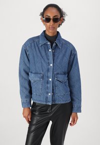 Quiltet jeansjakke i mellomblått med trykknapper, to frontlommer og en cropped design. Brukt over en svart turtleneck og svarte bukser.
