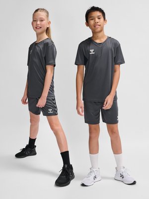 Twee kinderen staan naast elkaar in donkergrijze sportkleding en sneakers tegen een effen lichte achtergrond.