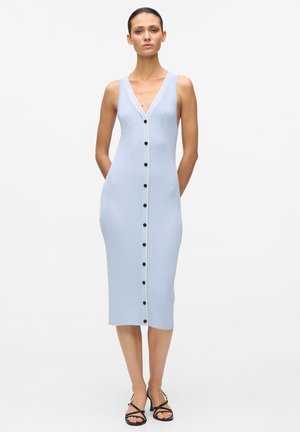 Naine, kes kannab varrukateta helesinist soonikkoes midi-kleit mustade nuppudega ees ning mustade rihmikuga sandaale, seisab käed selja taga.