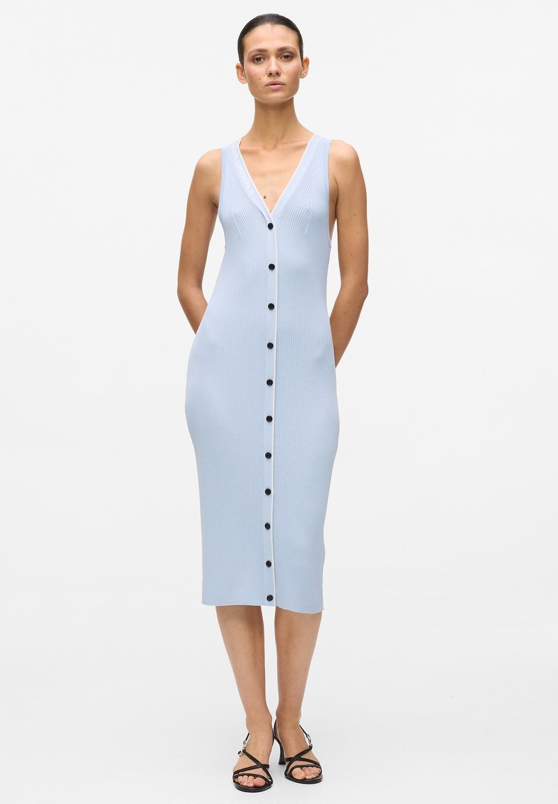 Femme portant une robe midi bleu clair sans manches en maille côtelée avec des boutons noirs sur le devant et des sandales noires à brides, debout avec les mains derrière le dos.