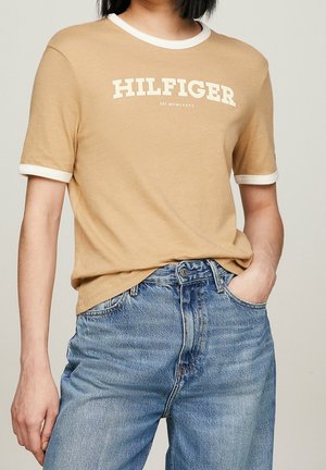 Béžové bavlněné tričko s bílým žebrovaným límcem a manžetami. Na hrudi je bílým tiskem logo "HILFIGER". Kombinováno s modrými džínami.