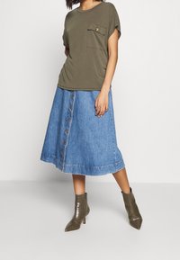 Olijfgroene T-shirt met zak, gecombineerd met een spijkerrok tot de knie met voorste knopen en een gebogen zoom. Olijfgroene enkellaarzen met textuurdetail.