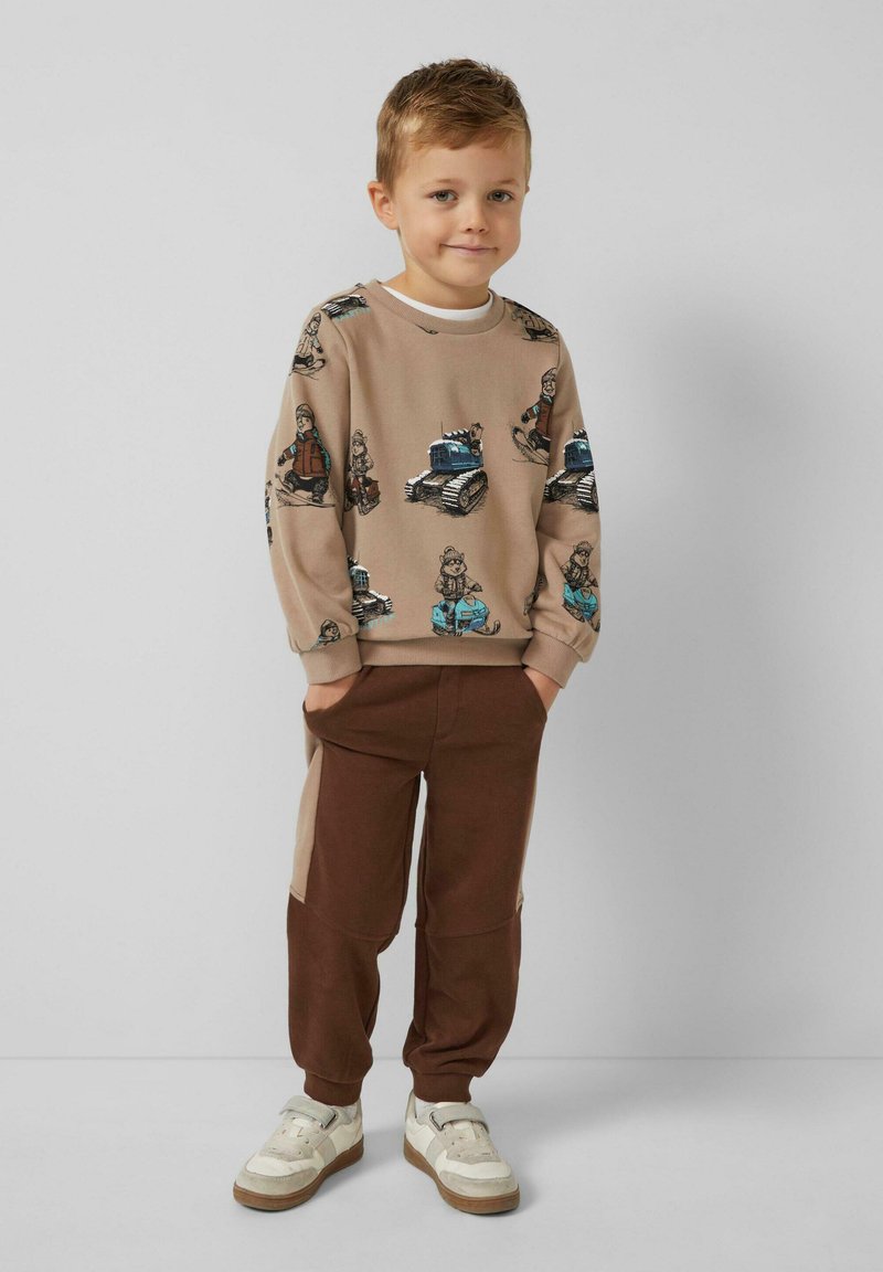 Beiger Pullover mit Cartoon-Mustern, braune Jogginghosen mit Seitenpaneelen und weiße Sneakers mit grauen Akzenten. Kind steht selbstbewusst da.