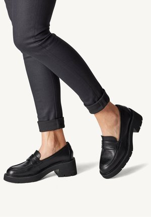 Sorte læder loafers med hævet hæl, der har en tekstureret overdel og stitch-detaljer, båret med mørke opfoldede bukser.