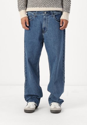 Personne portant un jean en denim bleu ample, des baskets blanches et grises, et un pull en tricot beige à motifs noirs.