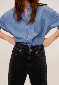 Chemise en denim bleu oversize avec une fermeture à boutons et une poche, accompagnée de pantalons noirs taille haute à taille élastique avec poches latérales.