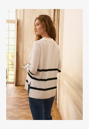 Witte gebreide trui met zwarte horizontale strepen, voorzien van een kraag en geribbelde zoom, gecombineerd met blauwe spijkerbroek.