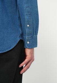 Denimová košeľa vo svetlomodrom odtieni, s ostrým golierom a gombíkovými manžetami. Tkanina má hladkú štruktúru a mierne vyblednuté miesta.