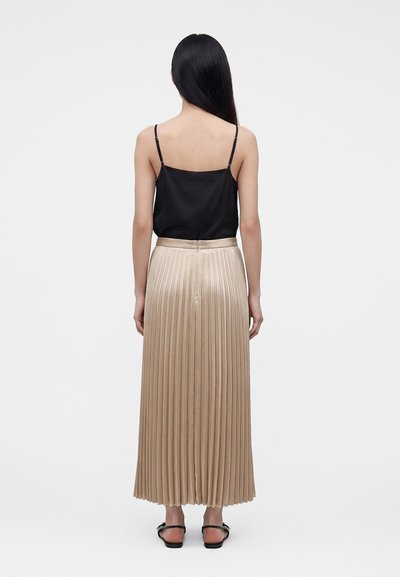 Femme aux longs cheveux noirs portant un haut noir à fines bretelles et une jupe midi plissée beige, debout de dos sur un fond uni.
