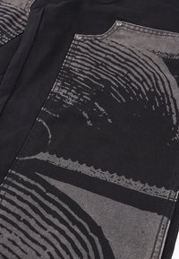 Κοντινό πλάνο μαύρου υφάσματος με ραμμένη τσέπη που φέρει αφηρημένα γκρι κυκλικά και κυματιστά γραφικά σε υφή υλικού.