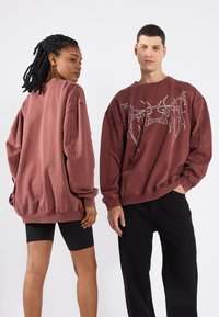 Sudaderas oversize en color marrón burdeos con diseño gráfico blanco, puños y dobladillos acanalados, combinadas con pantalones cortos y pantalones ajustados negros.