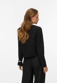 Crna skraćena jakna s dugim rukavima, kroja prilagođenog tijela, glatke teksture i srebrnih detalja na manšetama, uparen s crnim hlačama istog materijala.