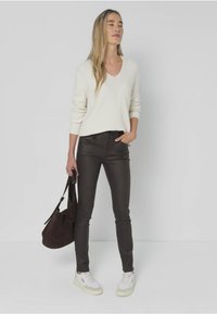 Jersey blanco de punto, pantalones ajustados negros, zapatillas blancas y bolso marrón. Pose de pie con fondo minimalista.