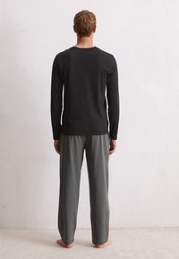 Man staat blootsvoets met zijn gezicht naar de muur, gekleed in een zwart long-sleeve shirt en zwart-wit geruite losse broek.