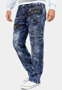 Pantaloni cargo in denim blu sbiadito con molteplici tasche, una finitura testurizzata e dettagli in pelle scura. Presentano chiusure con zip e bottoni.