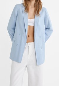 Femme portant un blazer bleu clair à double boutonnière sur un crop top blanc et un pantalon blanc taille haute, debout devant un fond blanc uni.