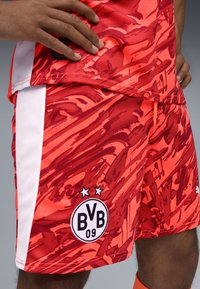 Shorts de football rouges avec un motif abstrait semblable à de la peinture, ornés d'une bande latérale blanche et d'un logo de club avec des étoiles. Le tissu texturé est visible.