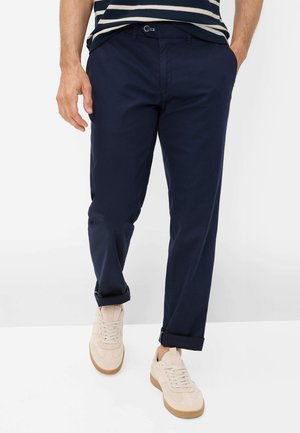 Mann trägt hochgekrempelte marineblaue Hose, beige Turnschuhe und ein marineblau-weiß gestreiftes Hemd, steht mit einer Hand in der Tasche.